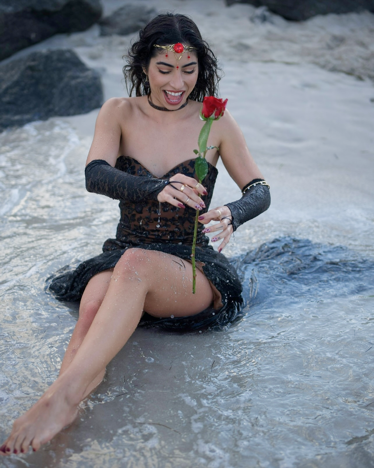 Selene Ashé sitting joyfully in the water at a beach, holding a red rose.